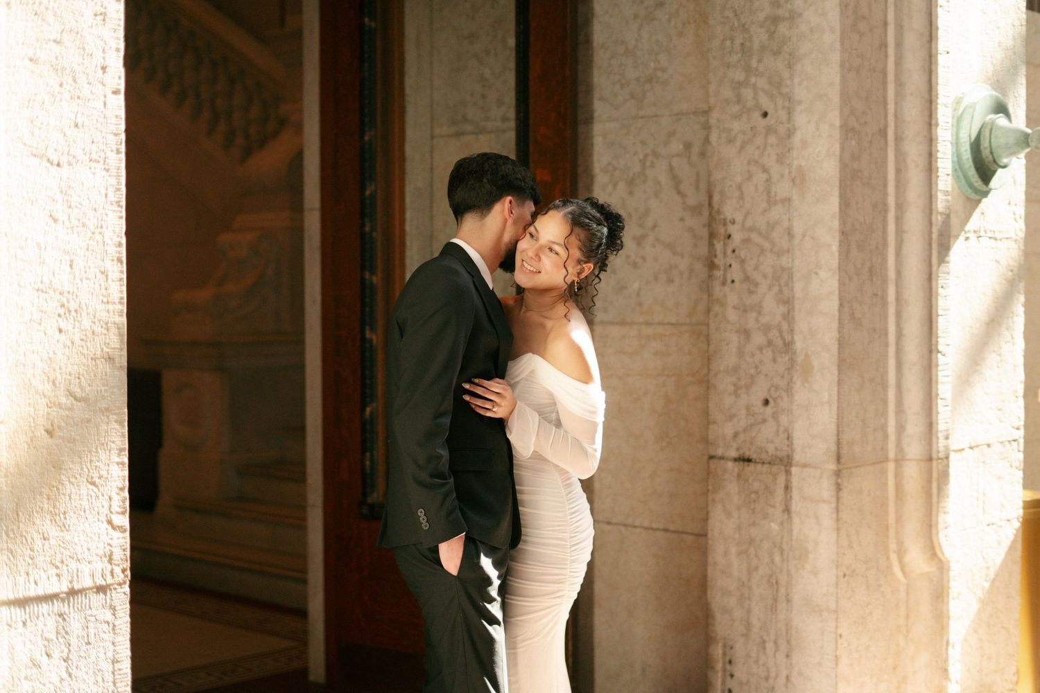 Couple embracing and kissing beside stone columns in warm sunlight.