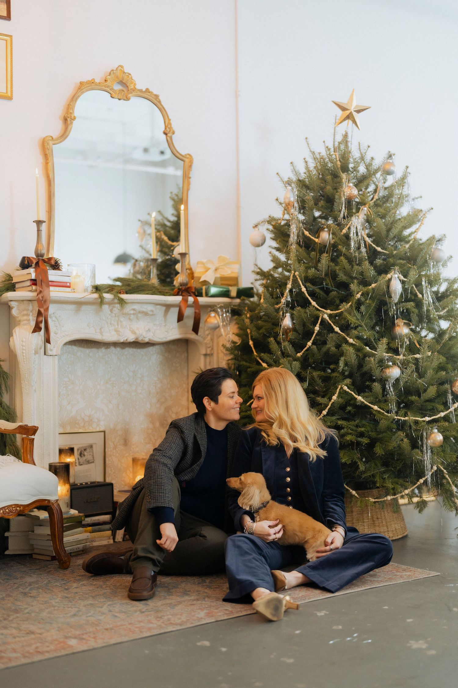 A couple sits together with their dog in front of a decorated Christmas tree in a cozy living room setting.