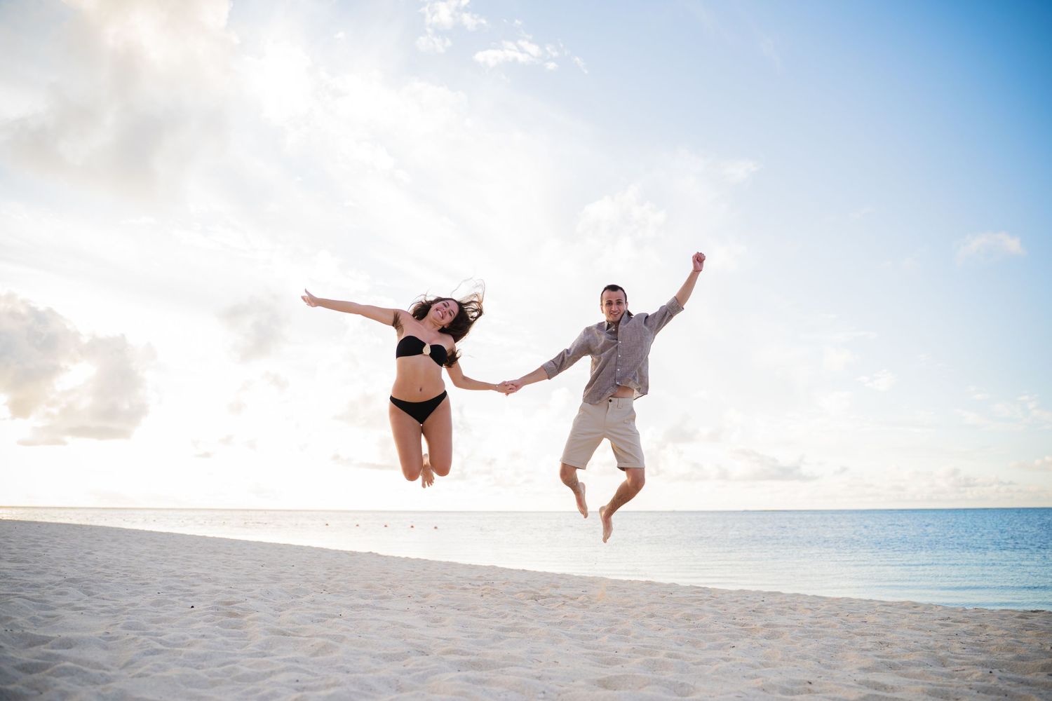 A couple jumps playfully on a tropical beach during sunset.
