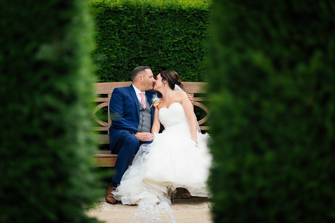 Couple in wedding attire sharing a romantic moment on a wooden bench in a lush garden setting.