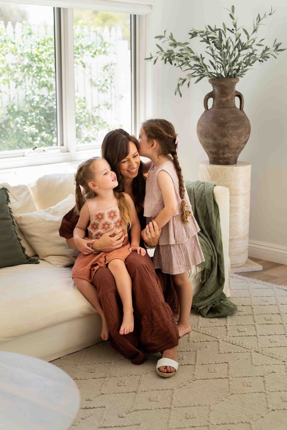 Mother sitting on a couch with her two children, one kissing her cheek while the other smiles during a Perth indoor family pop-up session