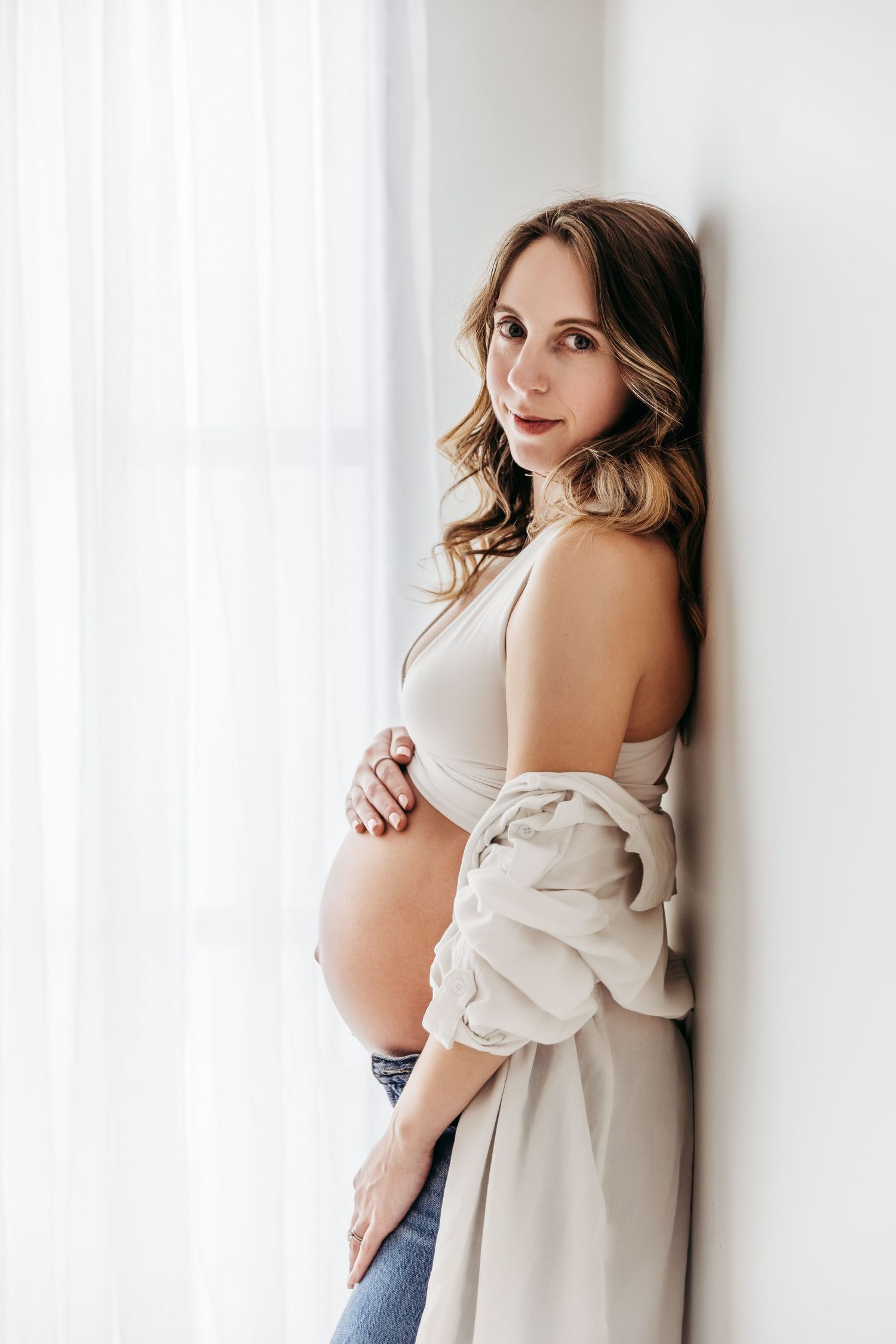 Expectant mother in white tank top and jeans poses against bright window with soft beige cardigan draped off shoulders.