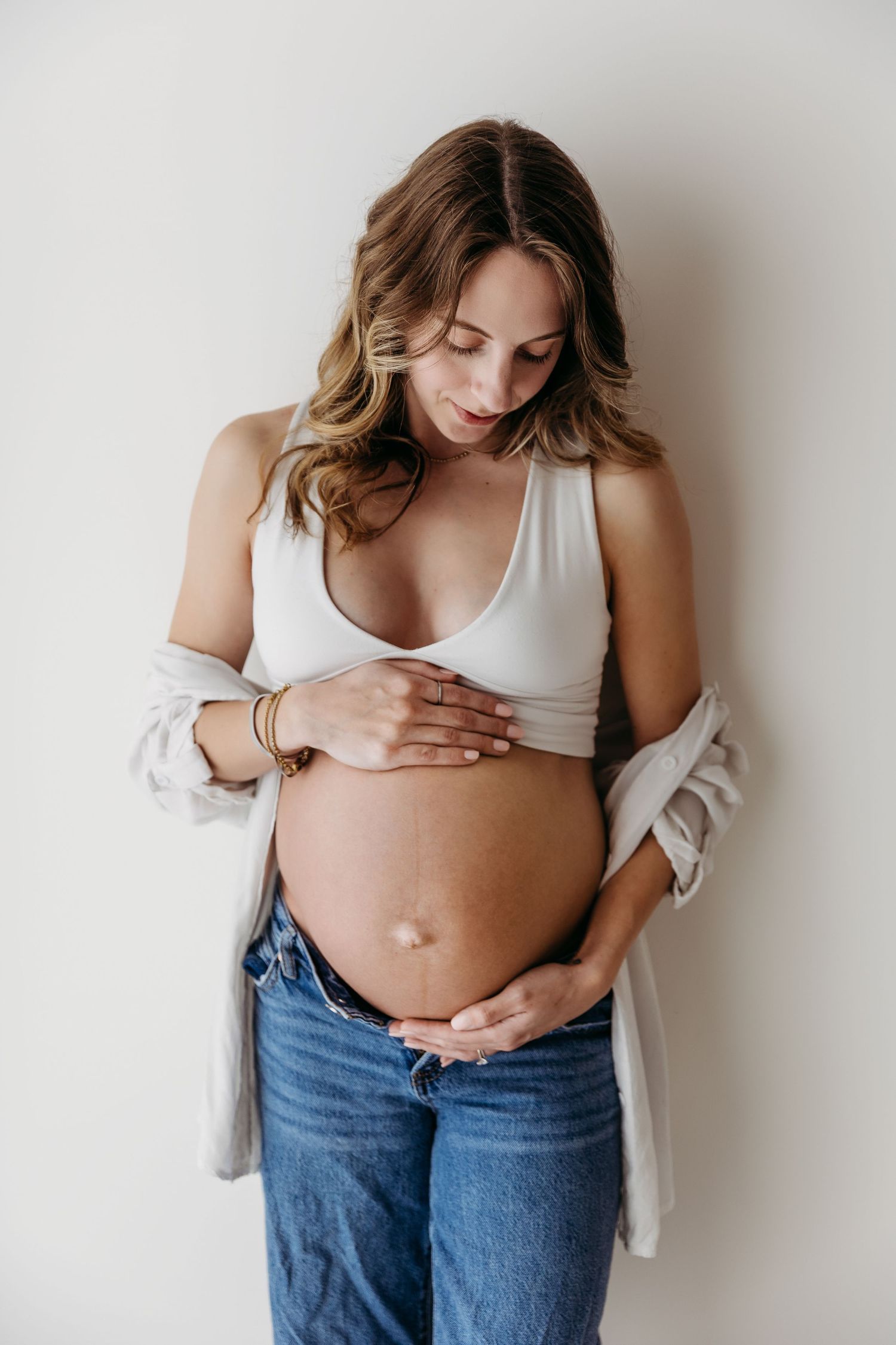 A pregnant woman in a white tank top and jeans cradles her baby bump while looking down with a gentle smile.