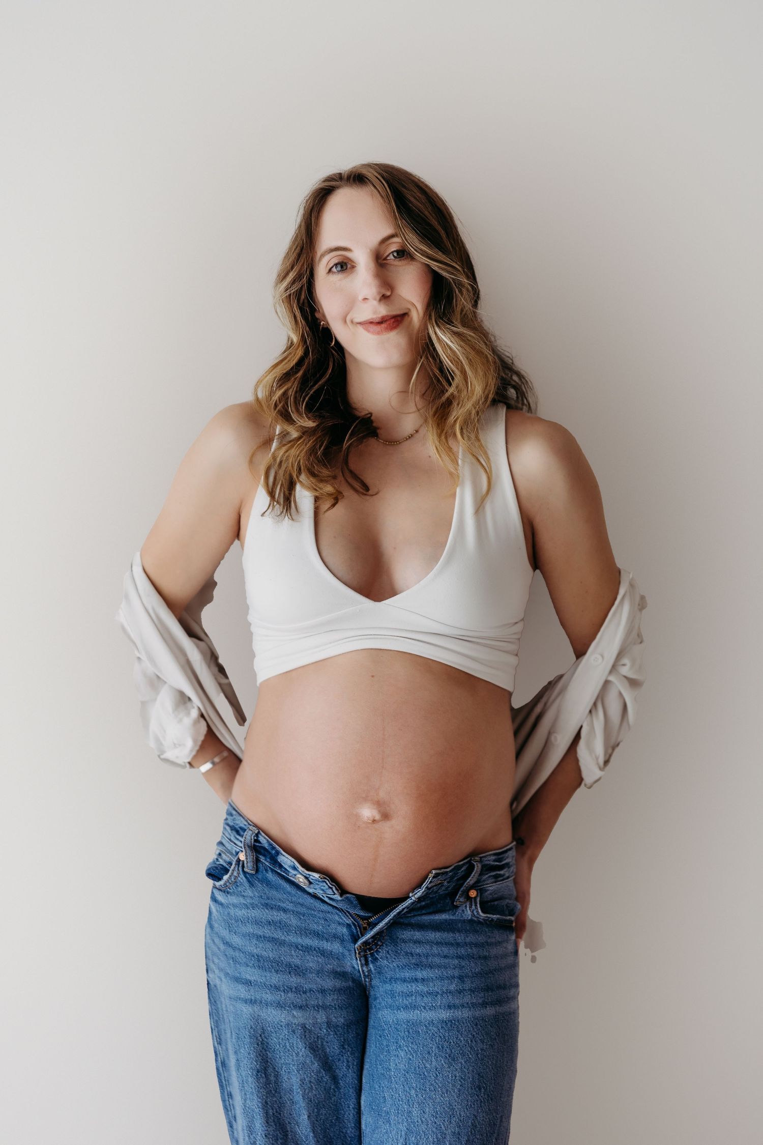 Expecting mother wearing white crop top and jeans poses against neutral wall showing growing baby bump.