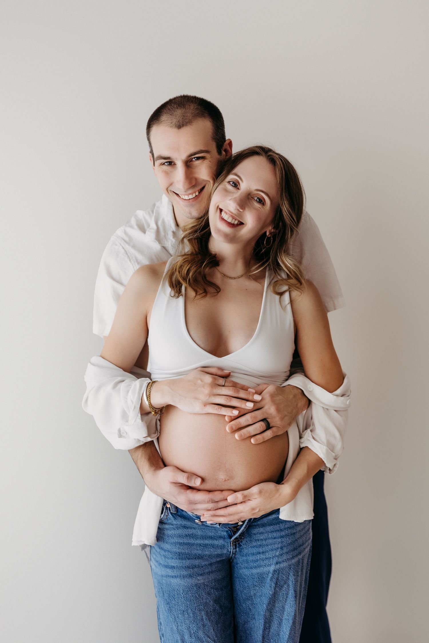 Maternity photo series showing a couple embracing against white wall, highlighting pregnant belly in jeans and white top.