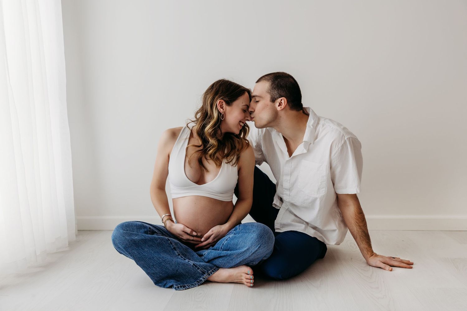 Couple in white shirts and jeans sitting close together on floor in bright minimalist room.