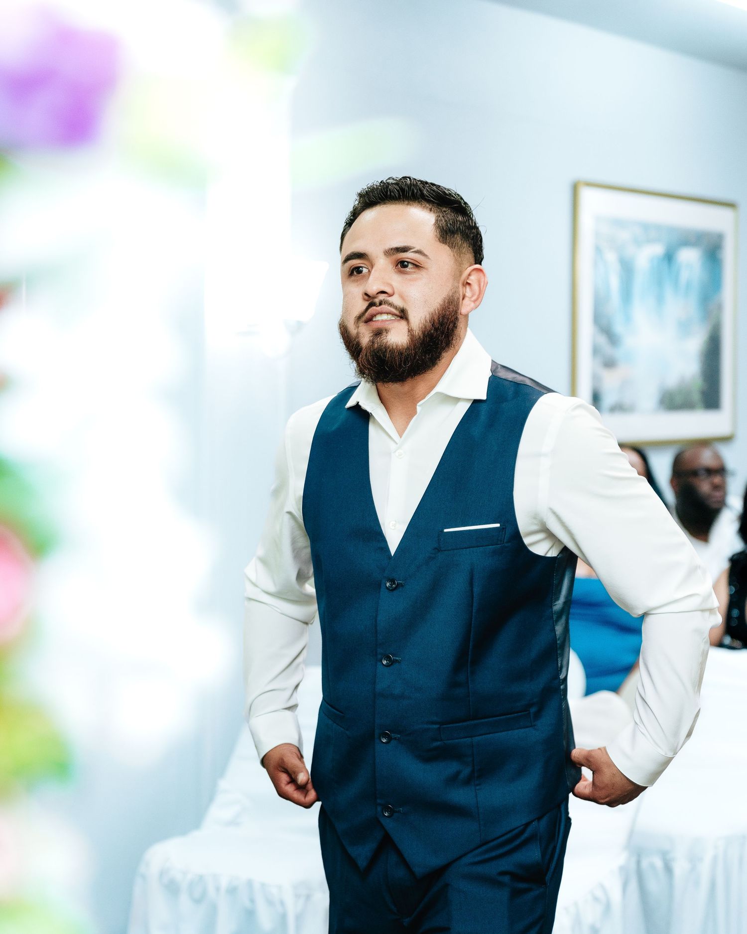 A person in a navy blue vest and white dress shirt poses confidently against a soft blurred background.