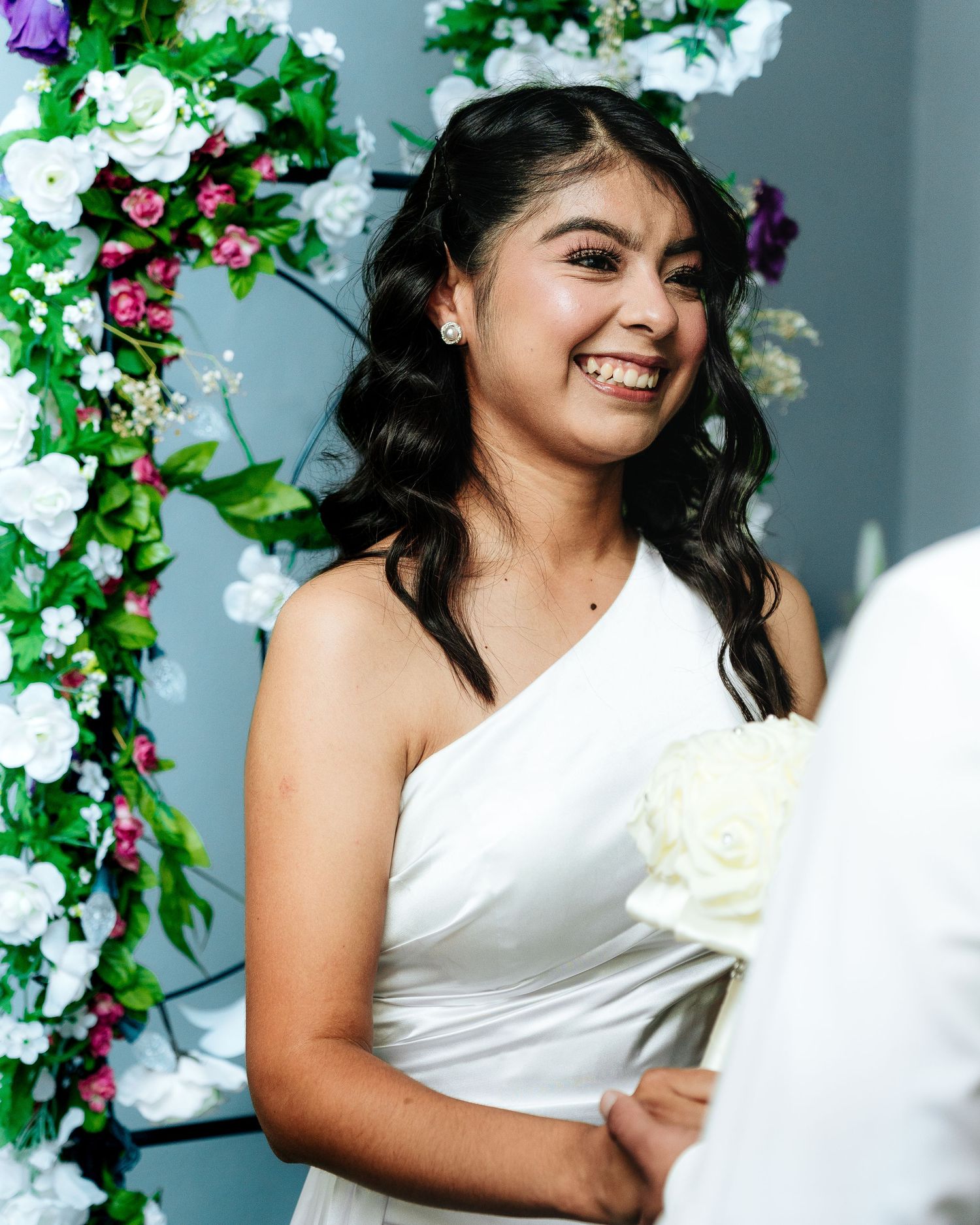 A joyful person in a one-shoulder white dress stands beside a floral arch with purple and white blooms.