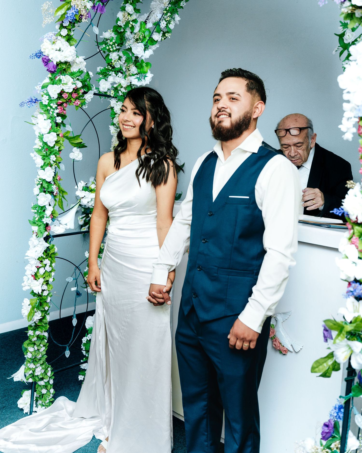 A wedding celebration under a floral arch with white and purple flowers, featuring a couple in formal attire.