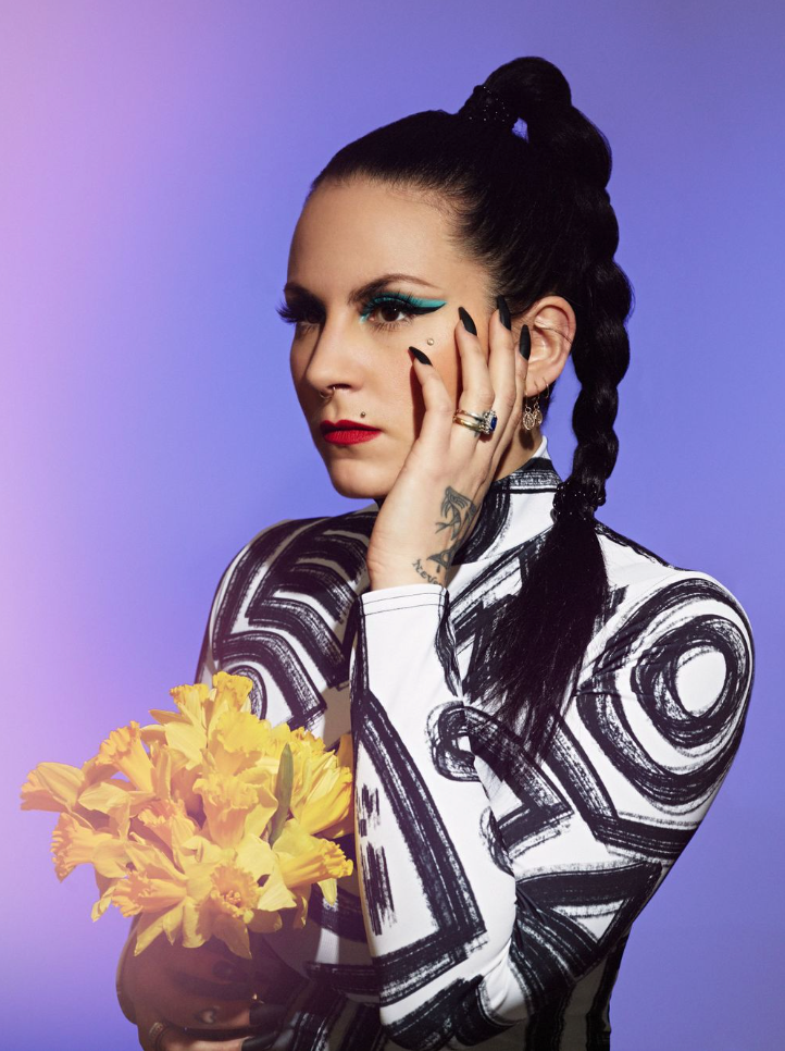 Artistic fashion portrait with dark hair, dramatic makeup, and yellow flowers against a purple background.