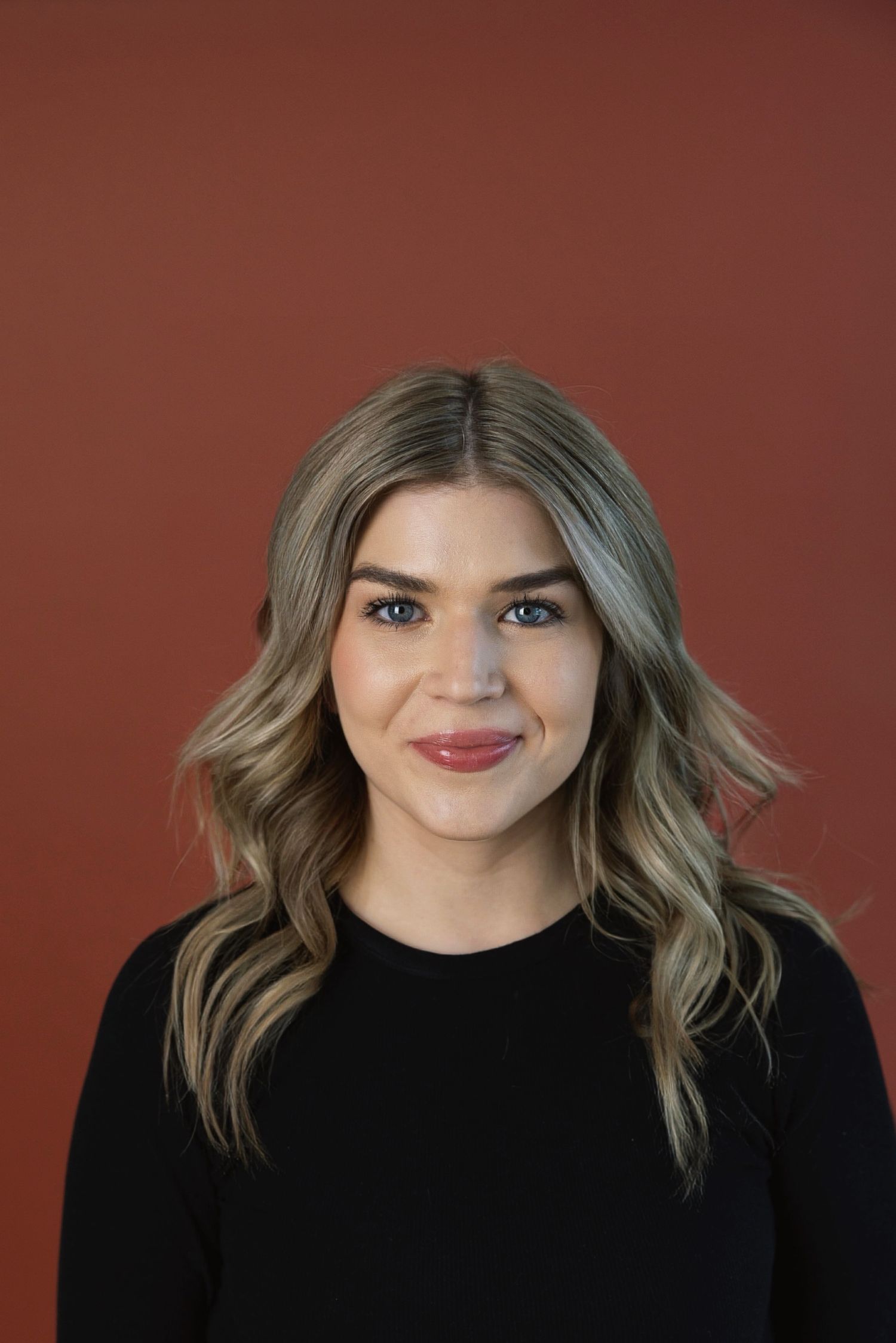 Young woman with highlighted wavy hair smiles in black top against a rich terracotta red studio backdrop.