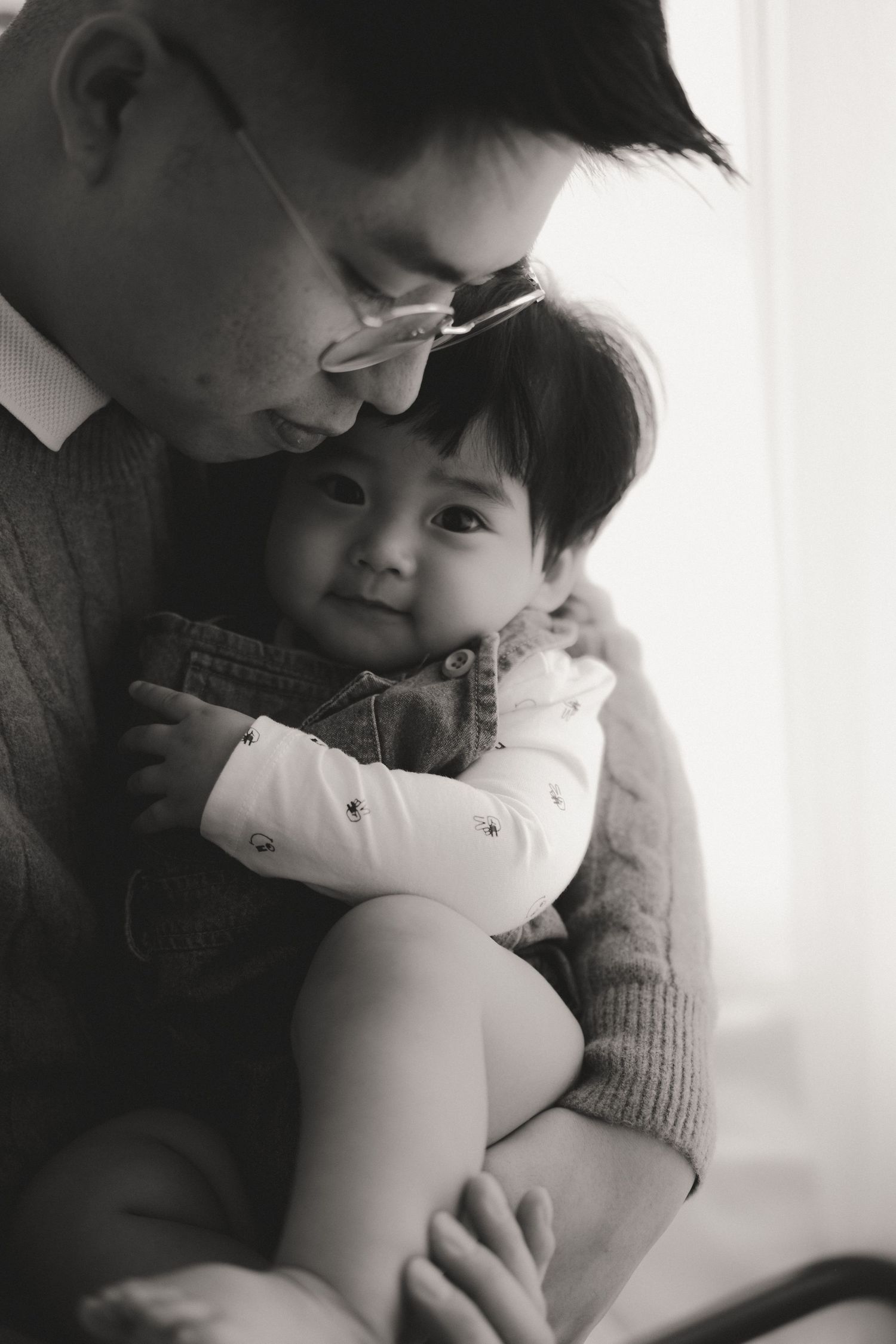 Black and white photo of a father tenderly holding his baby, sharing a sweet bonding moment near a bright window.一張黑白照片，照片中一位父親溫柔地抱著他的寶寶