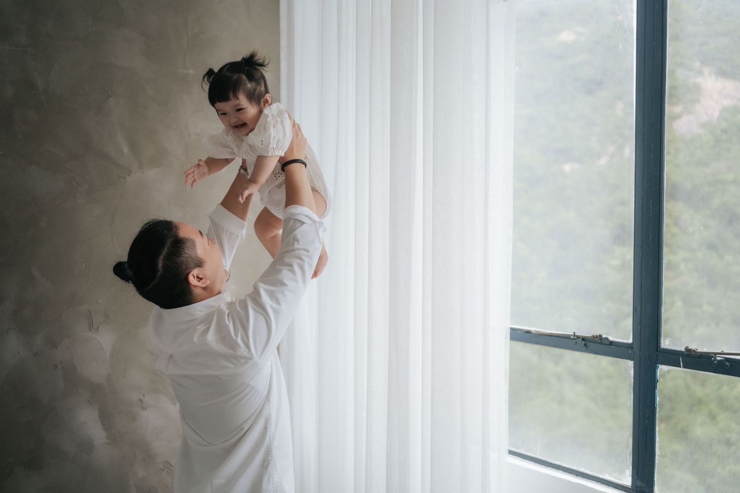 A mother joyfully lifts her baby in the air near a bright window with sheer curtains and a green view outside.一位母親高興地將她的寶寶舉到空中，窗外是明亮的窗戶，薄紗窗簾
