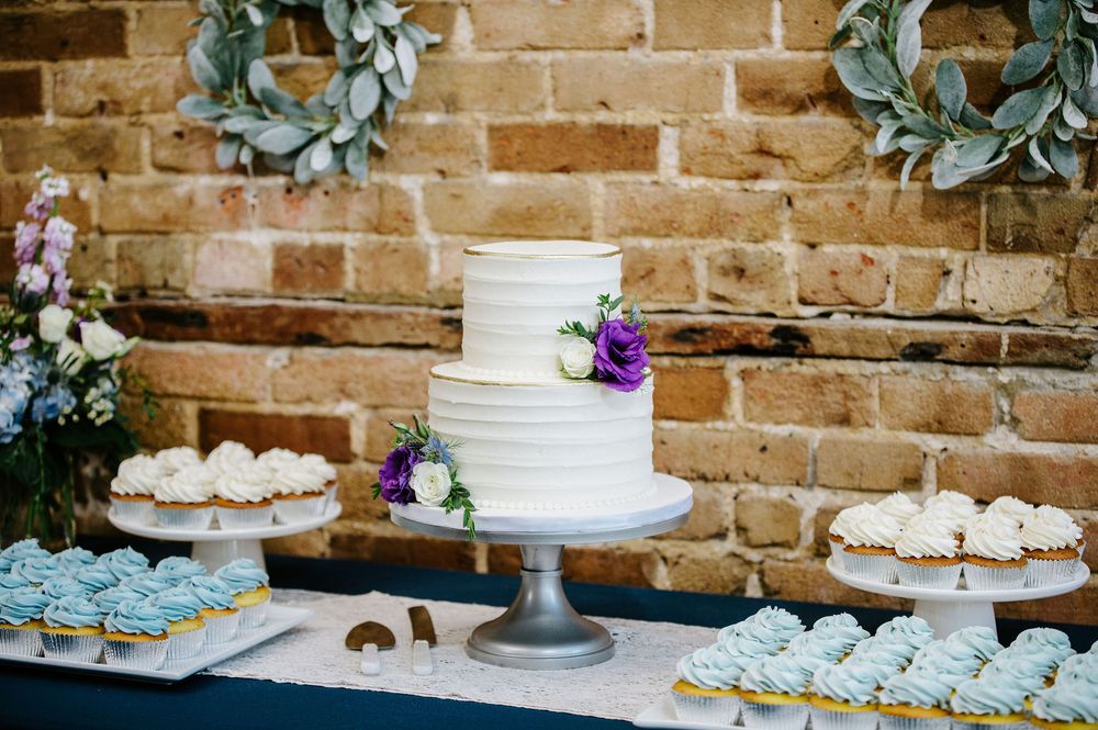 Wedding dessert display featuring white tiered cakes and cupcakes decorated with fresh flowers on rustic wooden tables.