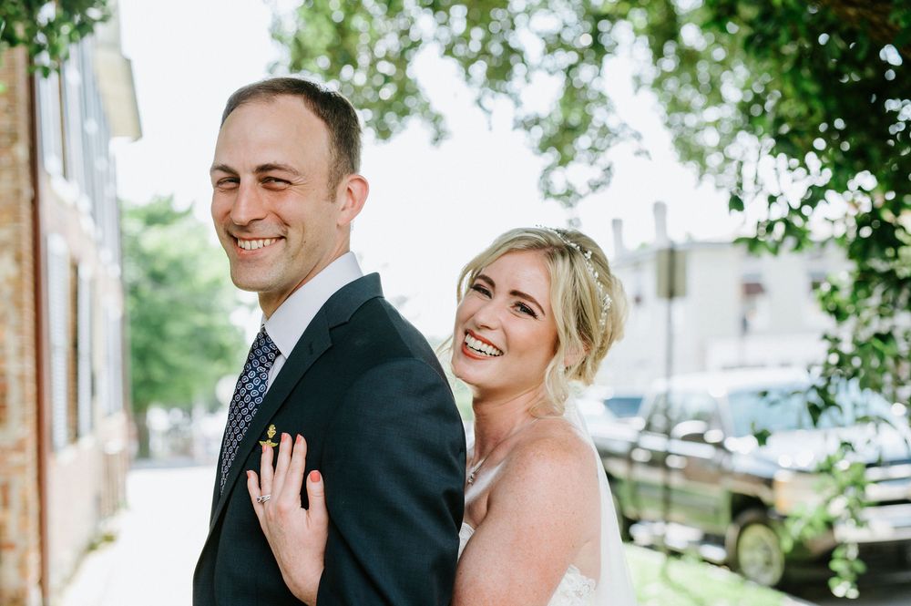 A joyful wedding couple shares a candid moment together outdoors on a sunny day with greenery in the background.