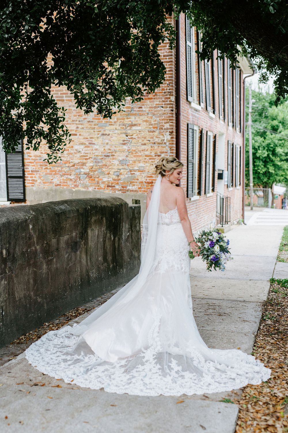 A stunning bridal gown with a long lace train photographed from behind on a brick-lined pathway.