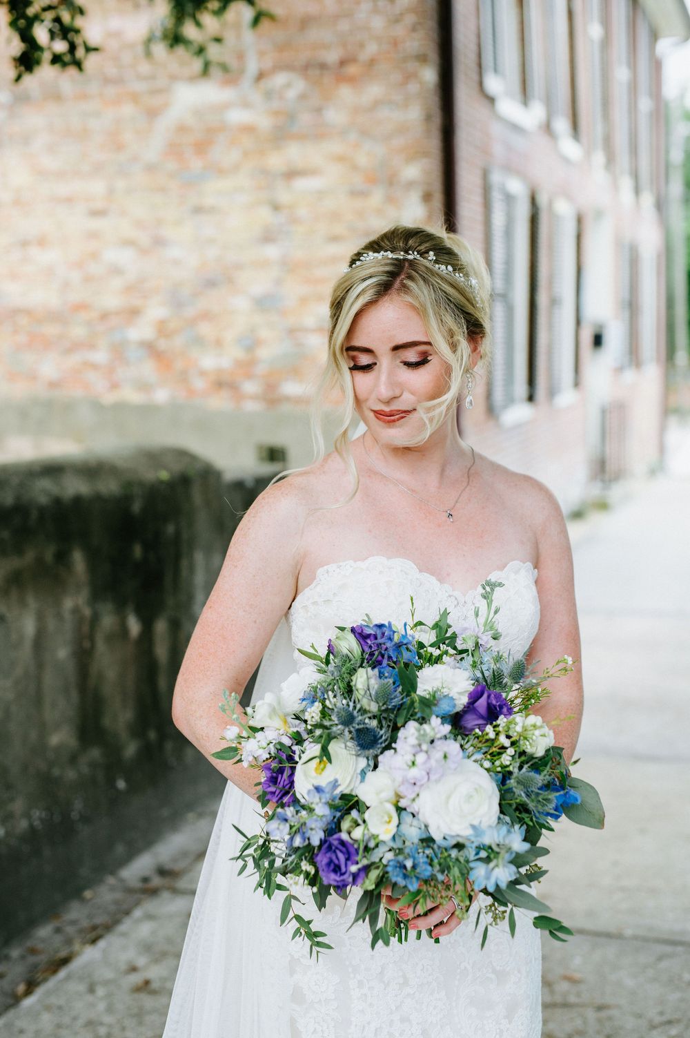 A bridal bouquet features stunning purple, blue and white flowers with lush greenery against a brick wall backdrop.