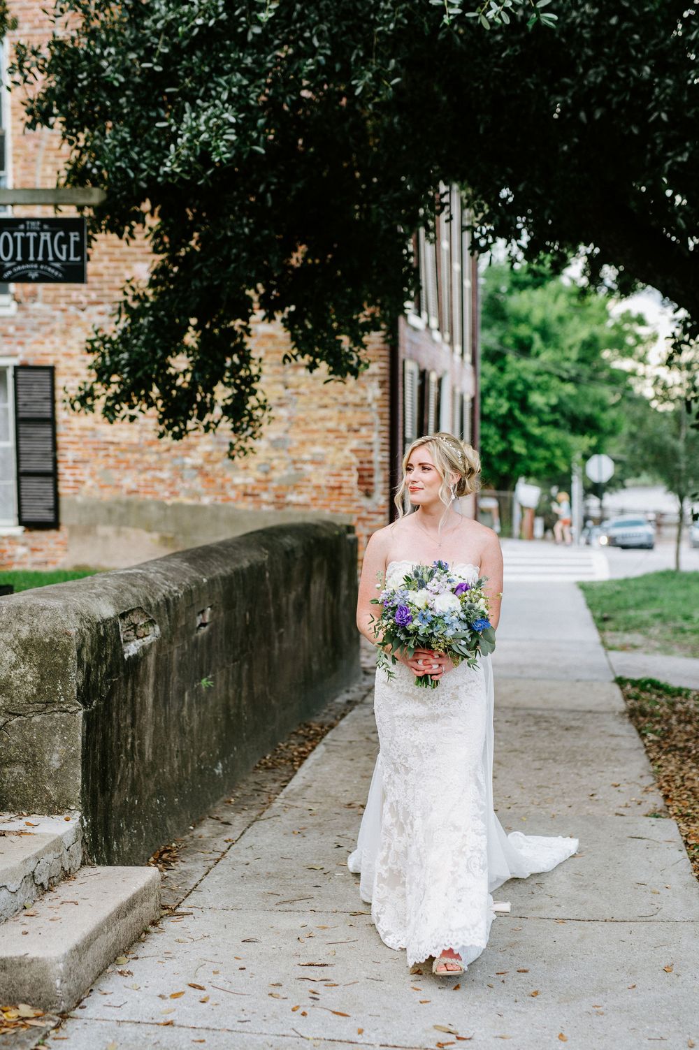 A bride in a white lace gown walks along a brick wall path holding a purple and white floral bouquet.