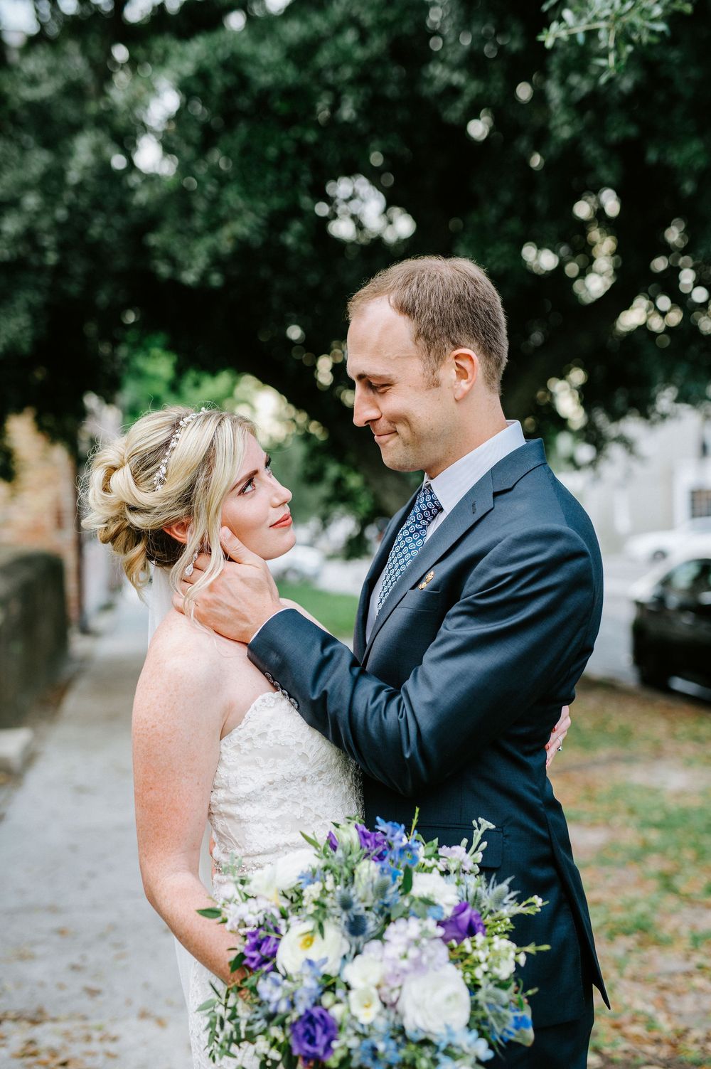 A romantic wedding couple embraces outdoors with a beautiful bouquet of purple and white flowers.