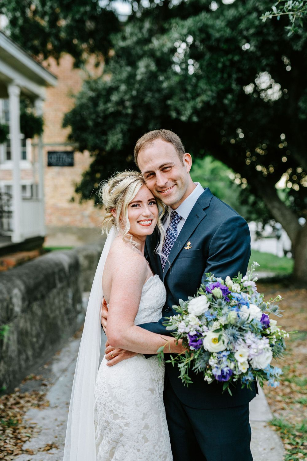 A joyful wedding couple shares intimate moments under trees while holding a vibrant purple and white bouquet.