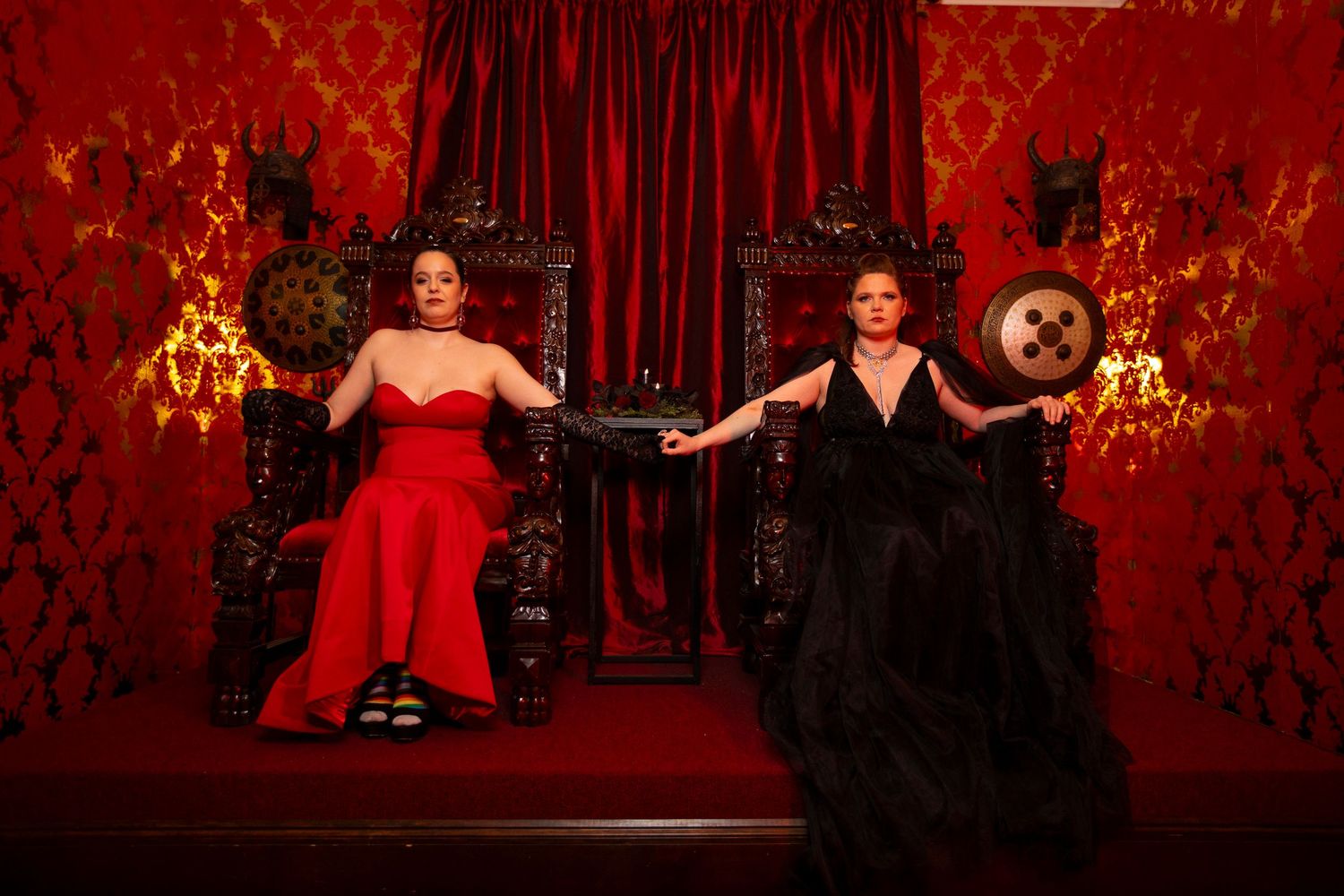 Two brides in red and black dresses sit beside each other and pose dramatically on stage in thrones for their wedding.