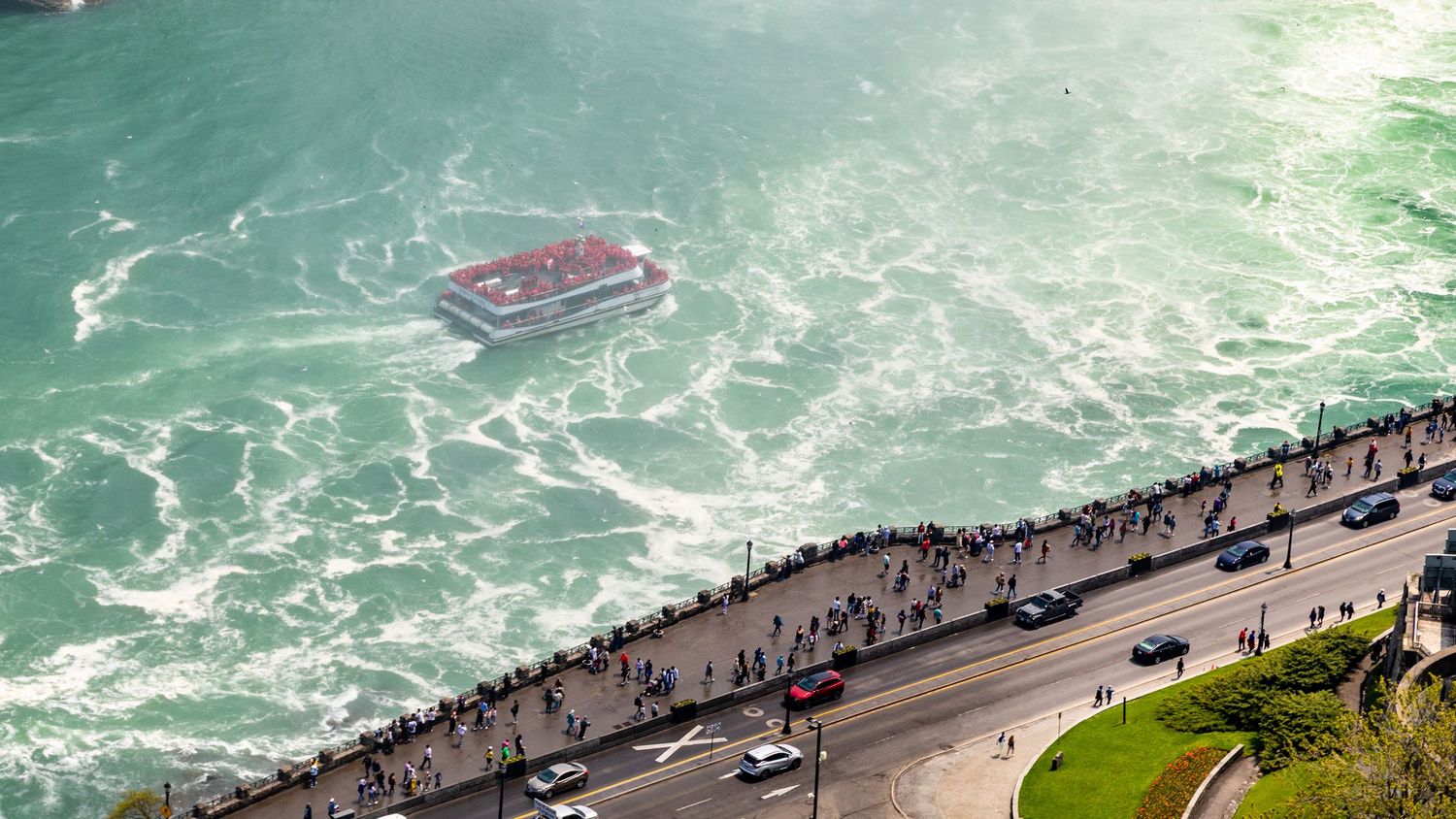 Aerial view of tourists gathered at Niagara Falls observation deck with a red boat navigating through turquoise waters.