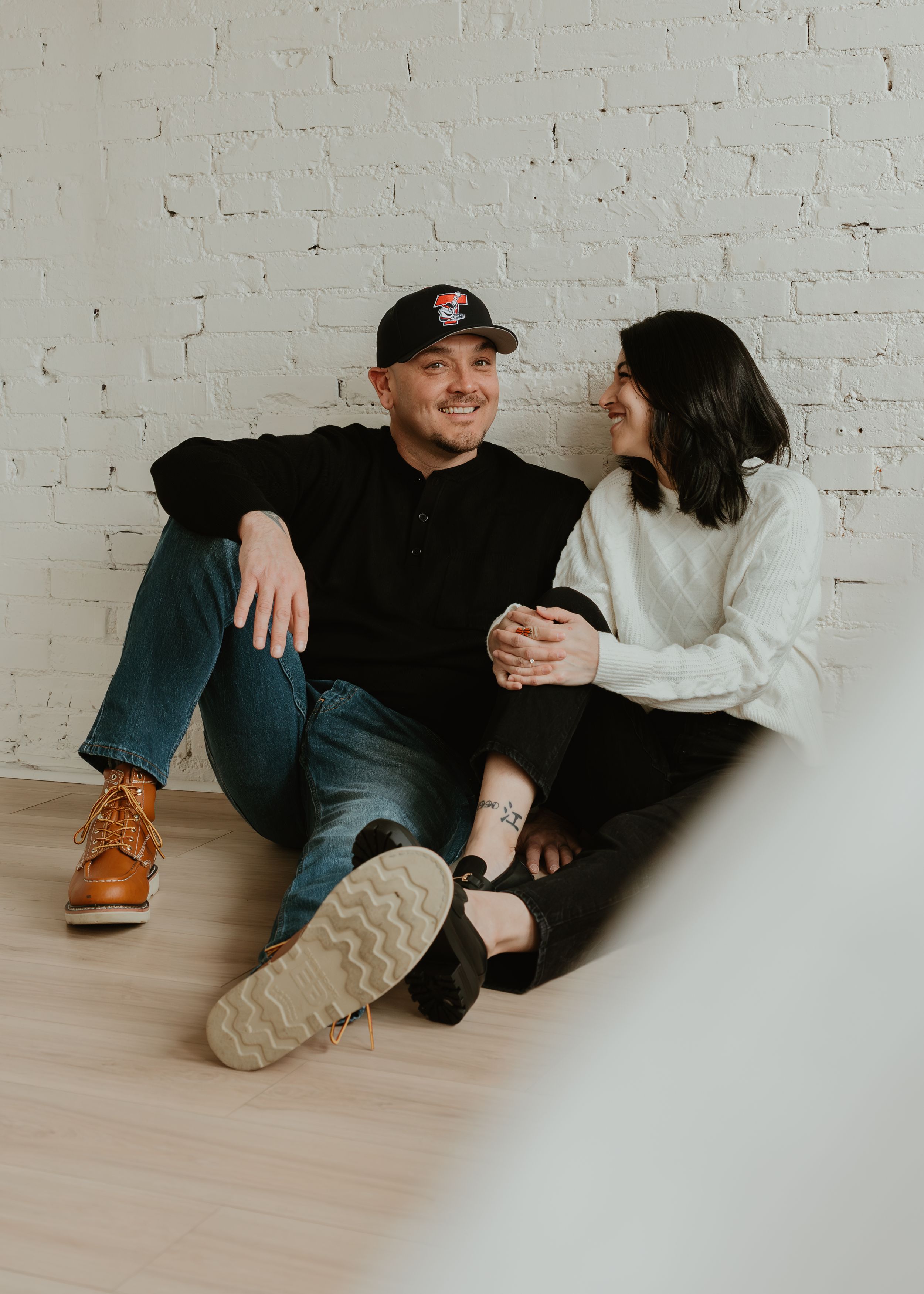 A couple relaxes together on a wooden floor in casual attire while taking candid photos.