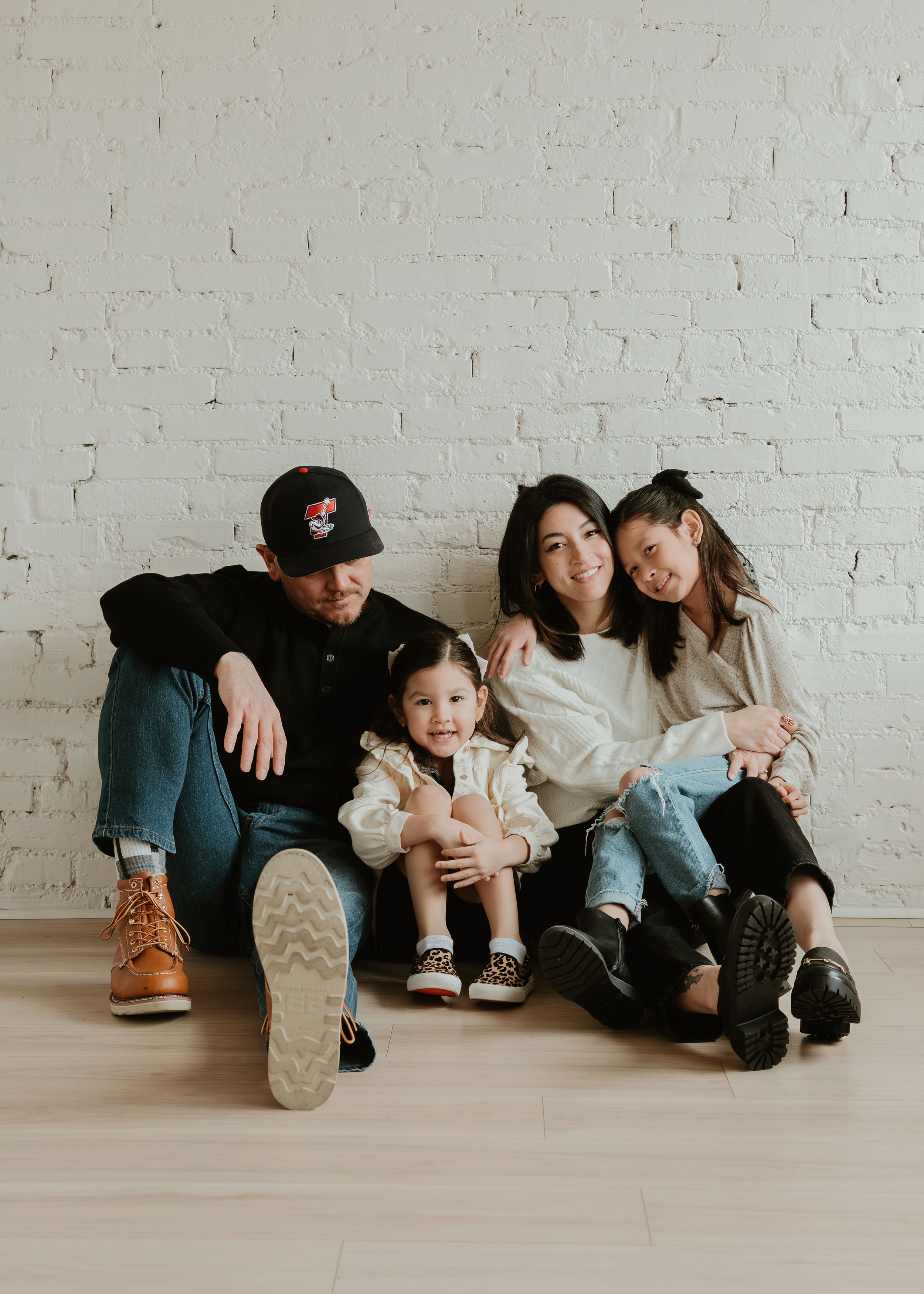 A family sits together on a white floor against a brick wall in a series of casual, connected poses.