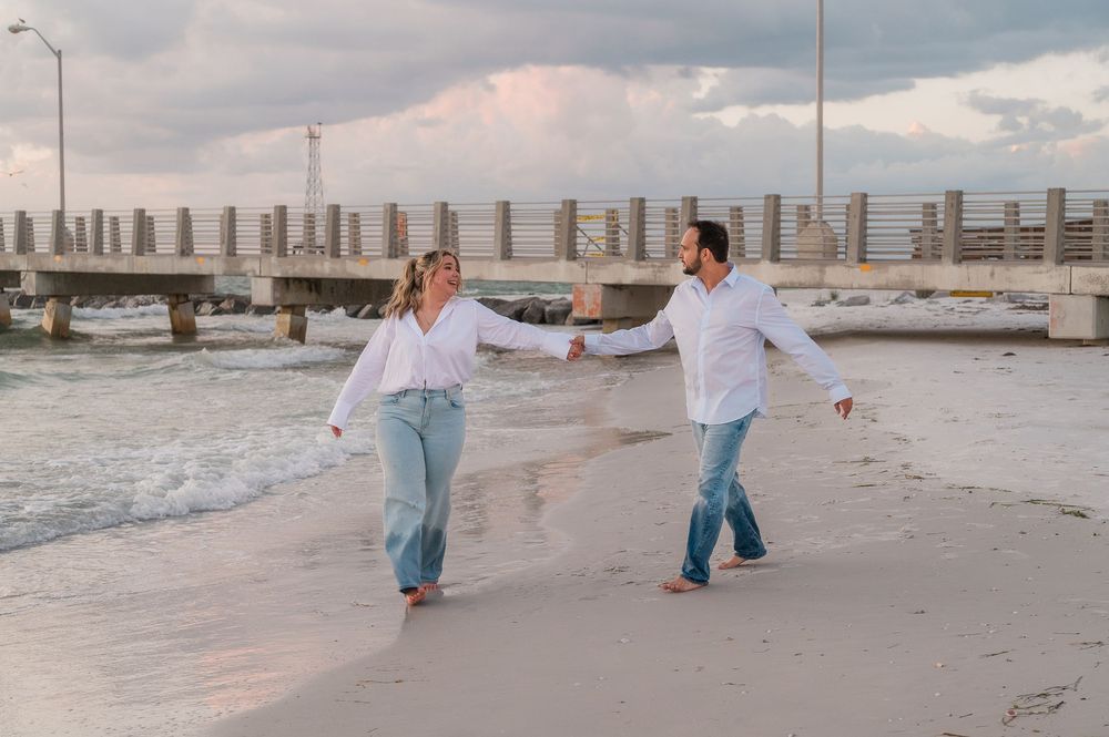 Two people in white tops dance joyfully together on a sandy beach at sunset.