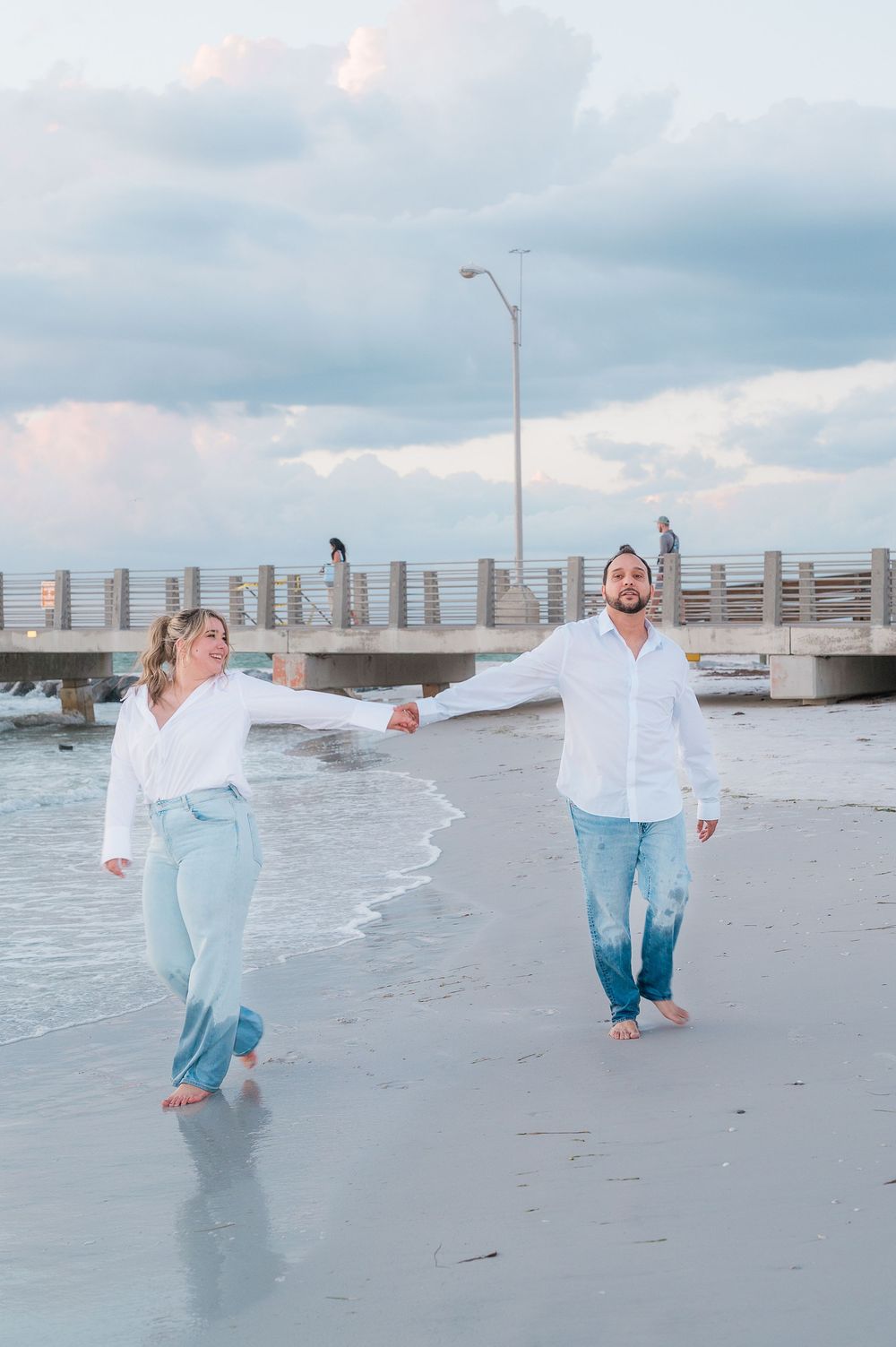 A couple in matching white shirts and jeans walk barefoot on a beach at sunset with a pier in the background.