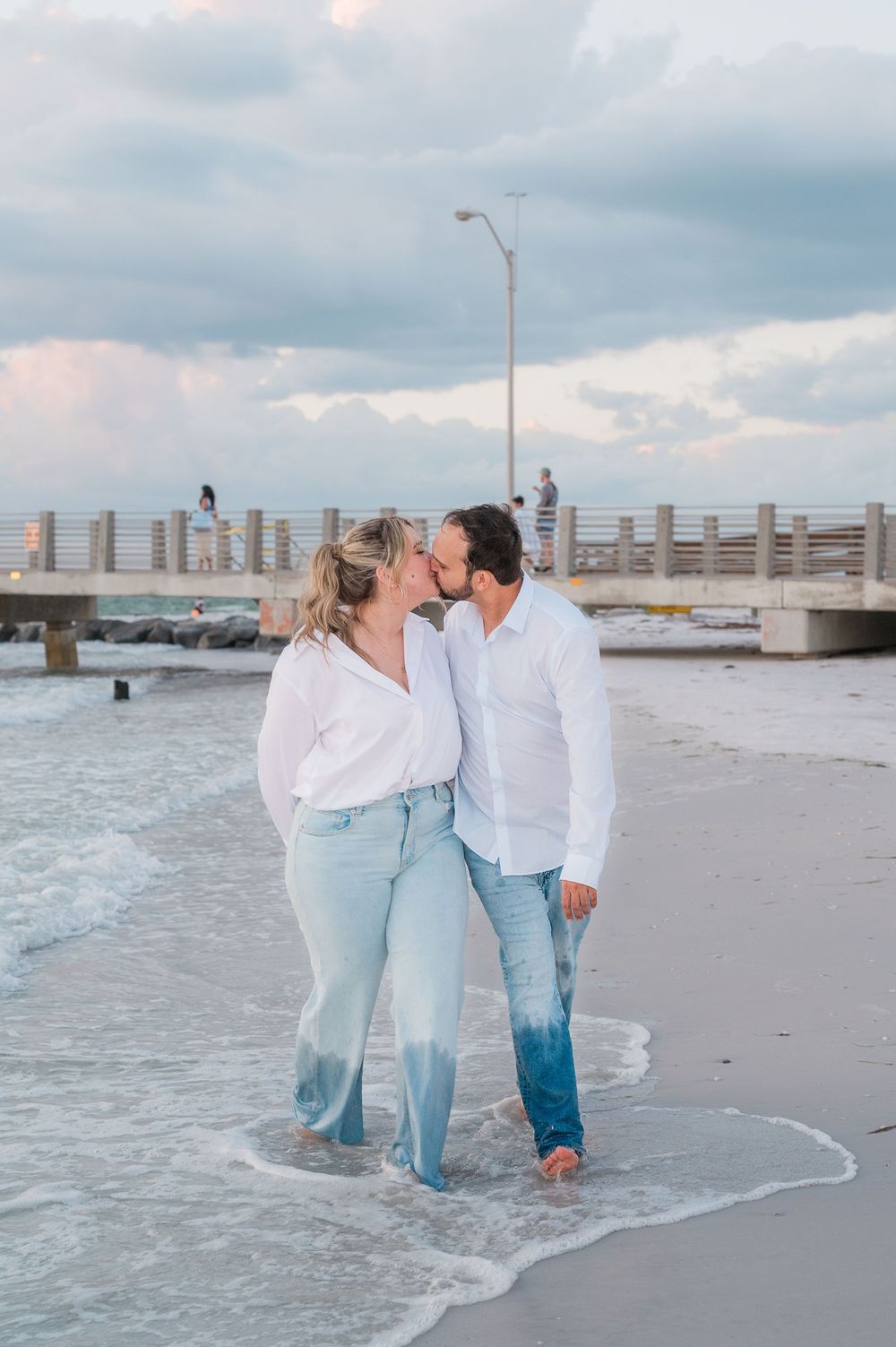 A romantic couple in matching light outfits shares an intimate moment while walking barefoot along the beach at sunset.