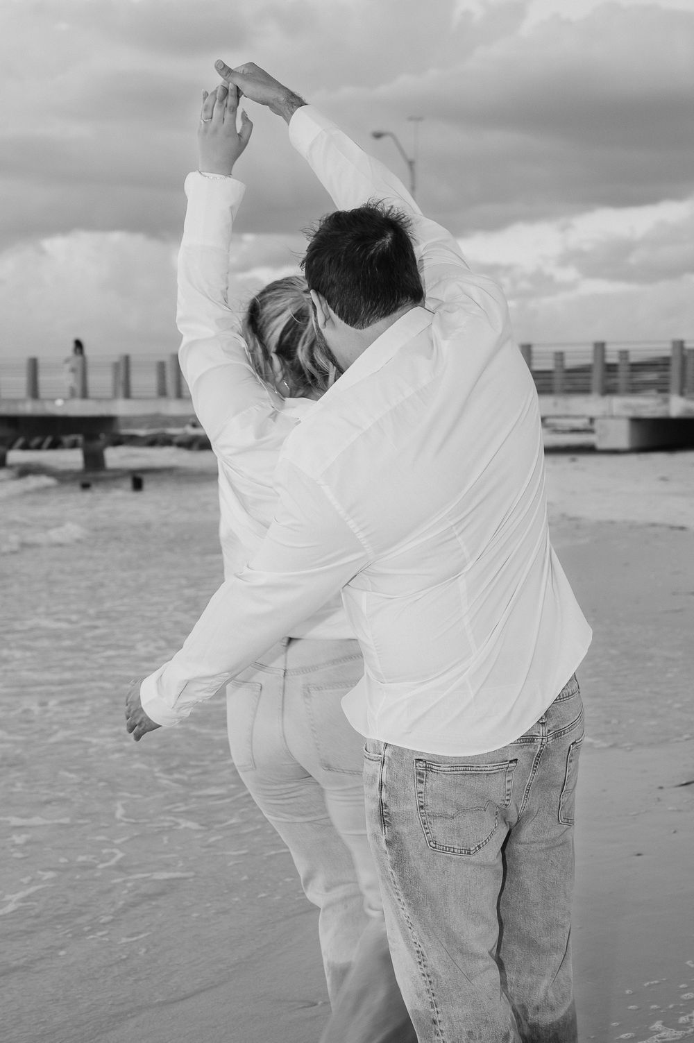 A couple dances together on a beach in white clothing against a cloudy sky and pier background.