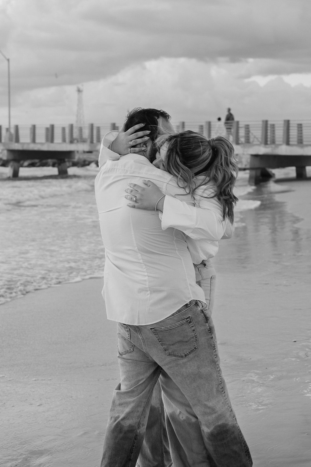 Playful couple in white shirts and jeans dancing together on a beach at sunset with city buildings in background.