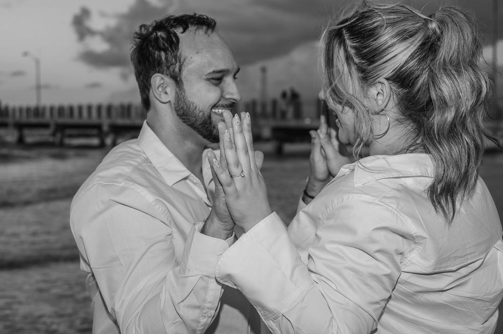 A happy couple sharing an emotional black and white moment on the beach with a pier in the background.