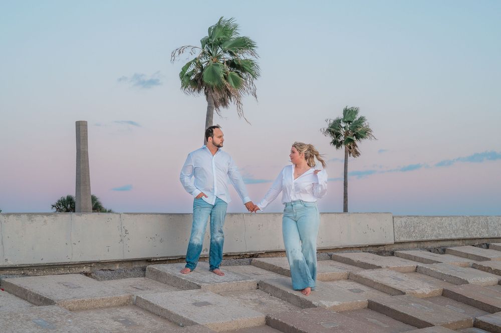 A couple in matching light blue outfits walks along concrete steps at sunset with palm trees in the background.