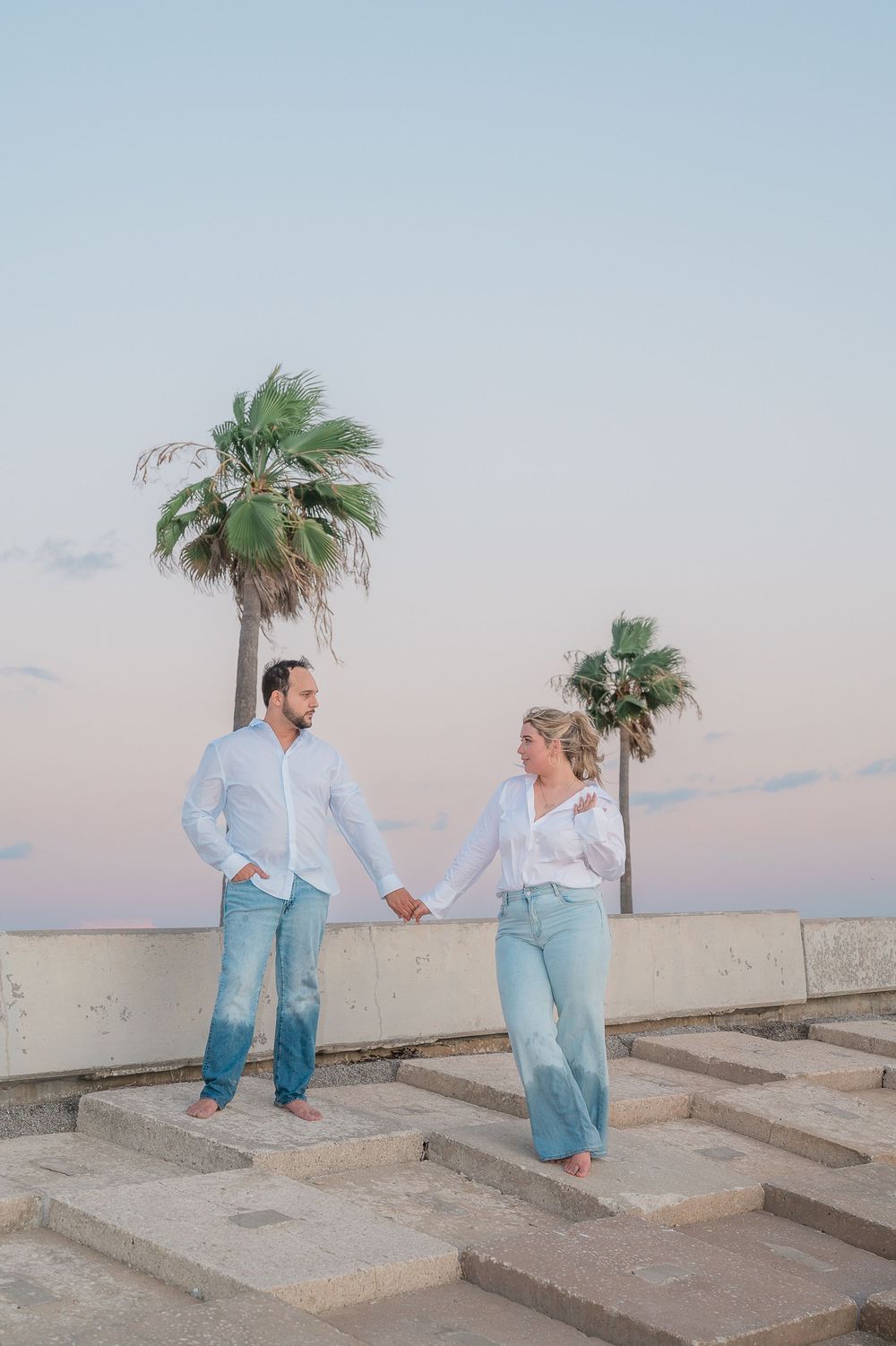 A couple in coordinating outfits poses on concrete steps with palm trees against a pastel sky.