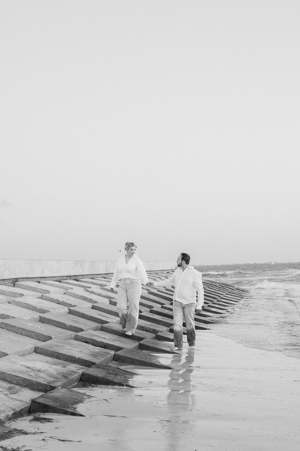 Couple walking along coastal jetty rocks in white outfits against a misty ocean backdrop.