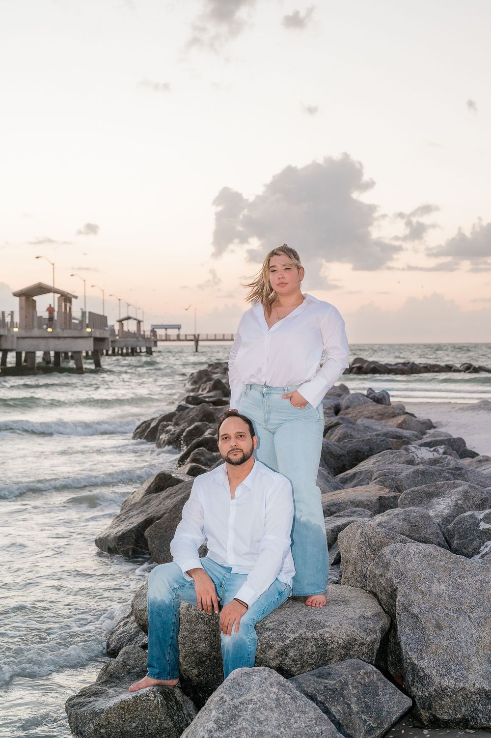 Couples portrait on rocky jetty at sunset with pier and ocean waves in background.