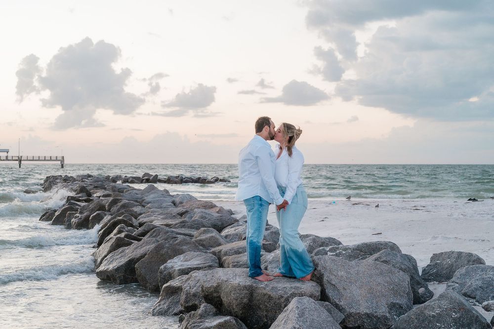 Couple in white and blue outfits embracing on rocky beach during sunset.