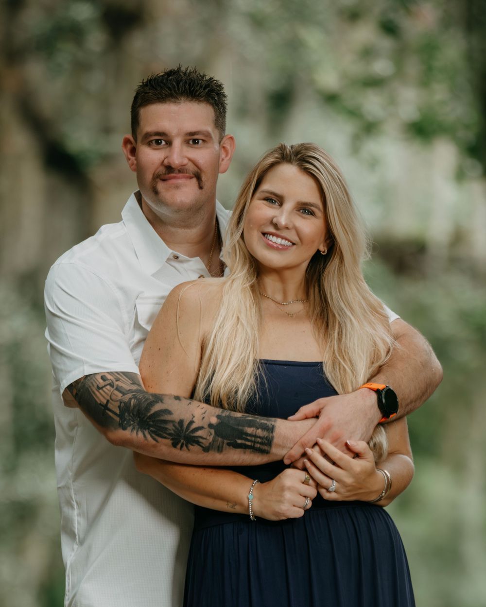 A smiling couple poses outdoors, man in white shirt with tattoos embracing blonde woman in navy dress.