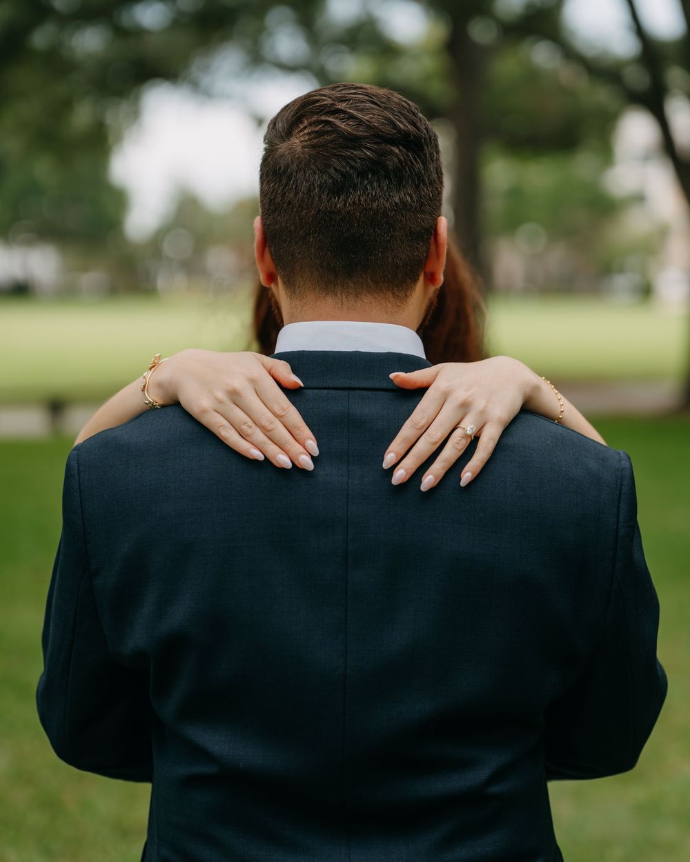 A groom in a navy suit seen from behind with a bride's hands resting on his shoulders, wearing an engagement ring.