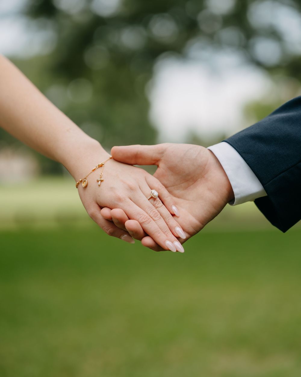 Couple holding hands outdoors, woman wearing gold bracelet and ring, man in navy suit, green blurred background.