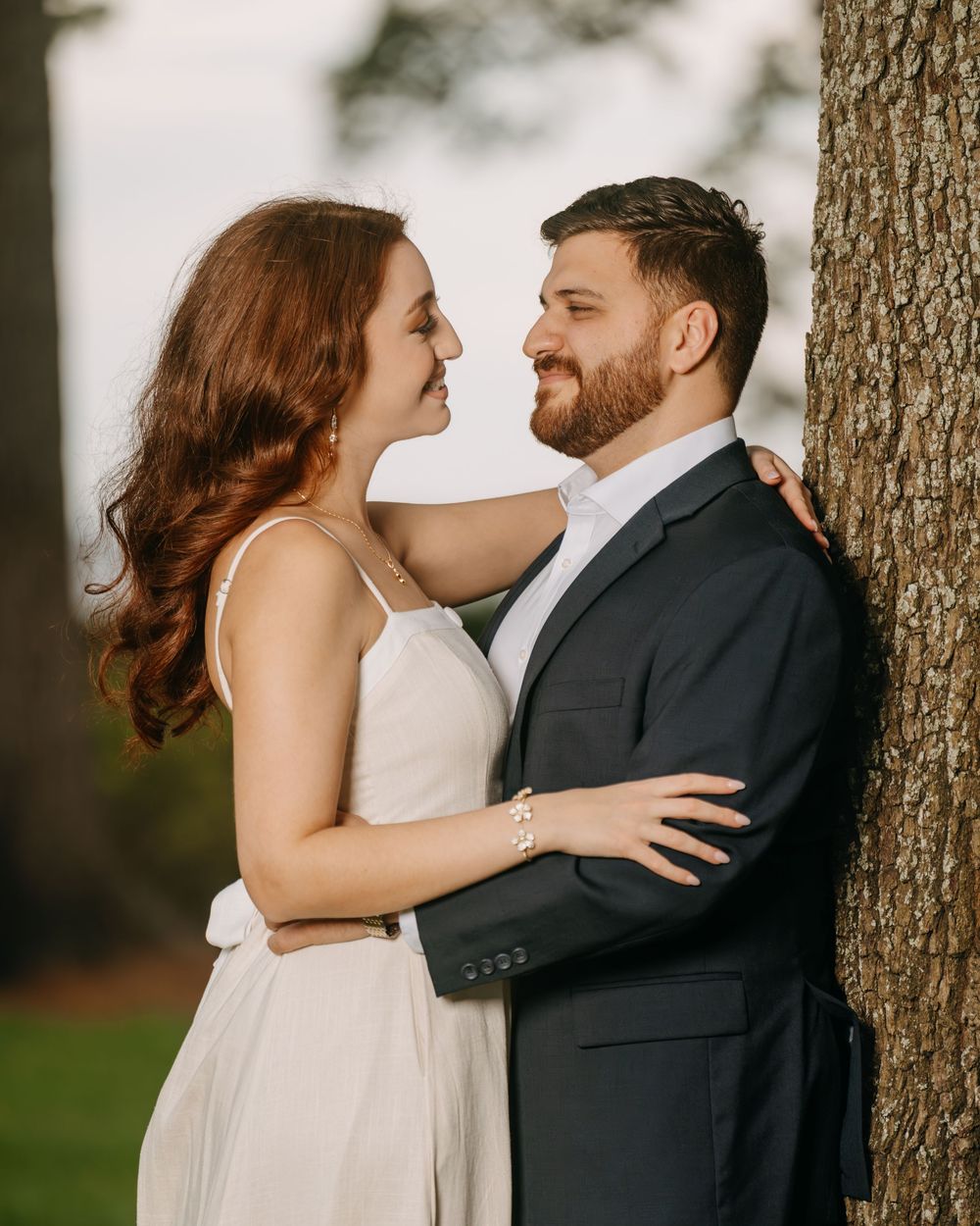 A couple in formal attire embraces near a tree, smiling at each other during an outdoor engagement photo session.