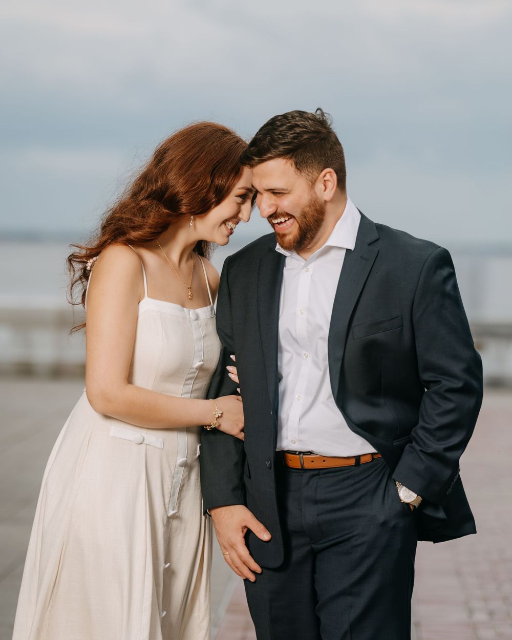 A smiling couple in formal attire share a romantic moment on a waterfront promenade, touching foreheads affectionately.