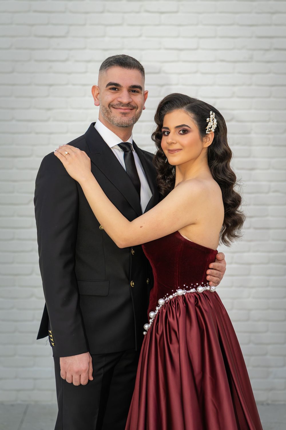 A couple poses together against a white brick wall, with her wearing a burgundy gown and him in a black suit.