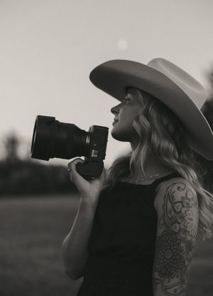 Black and white portrait of photographer with tattoo and wide-brim hat holding camera.