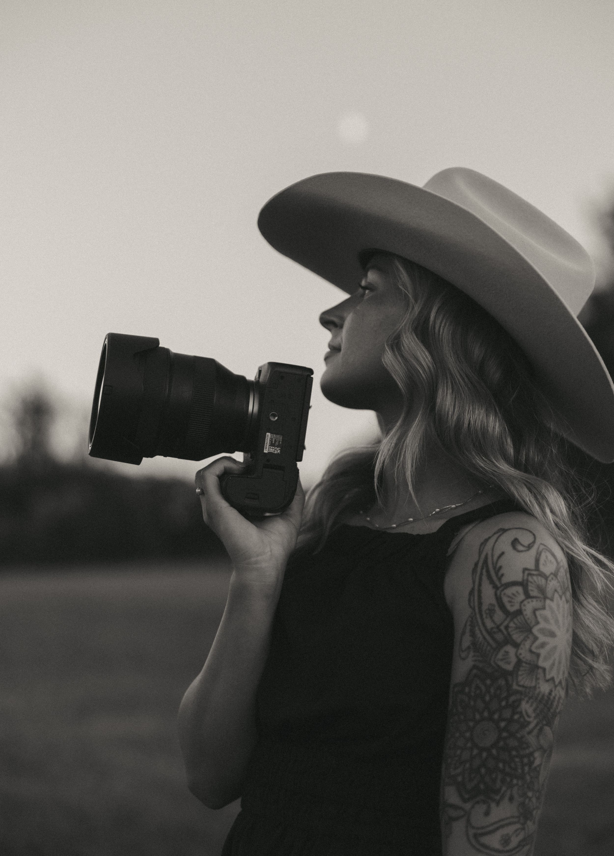Black and white portrait of photographer with tattoo and wide-brim hat holding camera.