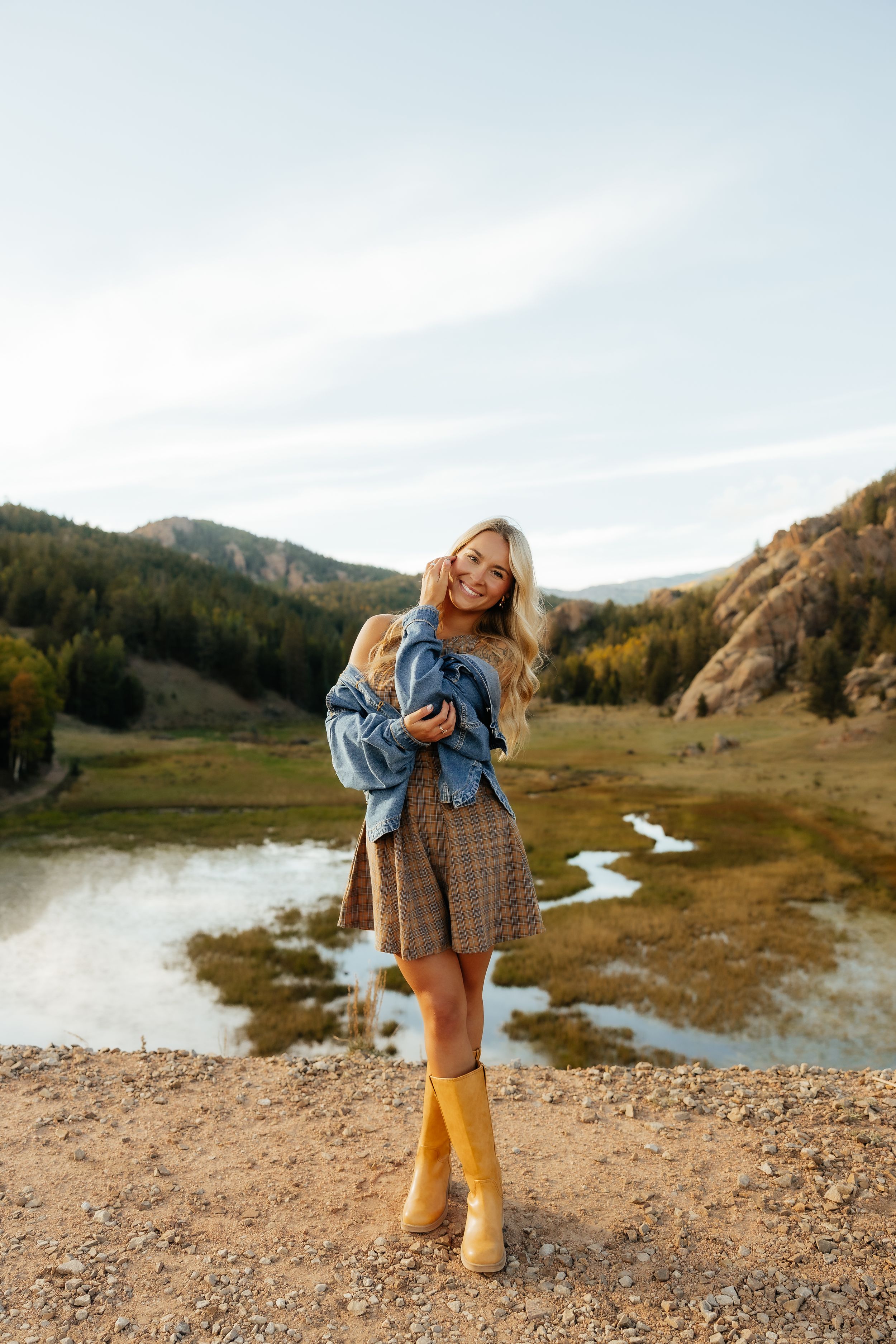 Smiling blonde woman in plaid skirt, yellow rain boots, and denim jacket posing near a mountain lake in autumn.