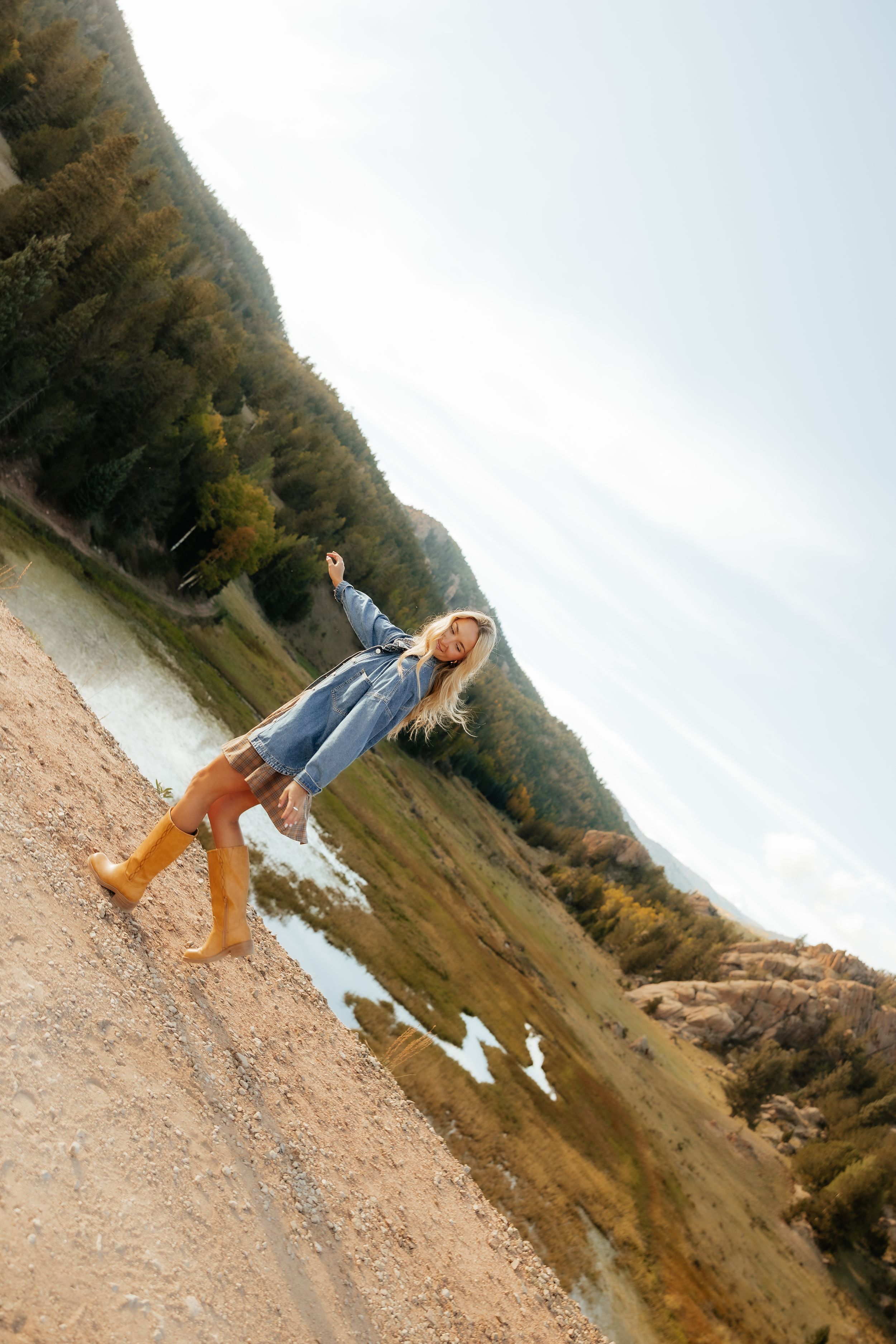 Young blonde woman in denim jacket and orange rain boots posing on a hillside with a scenic mountain lake backdrop.