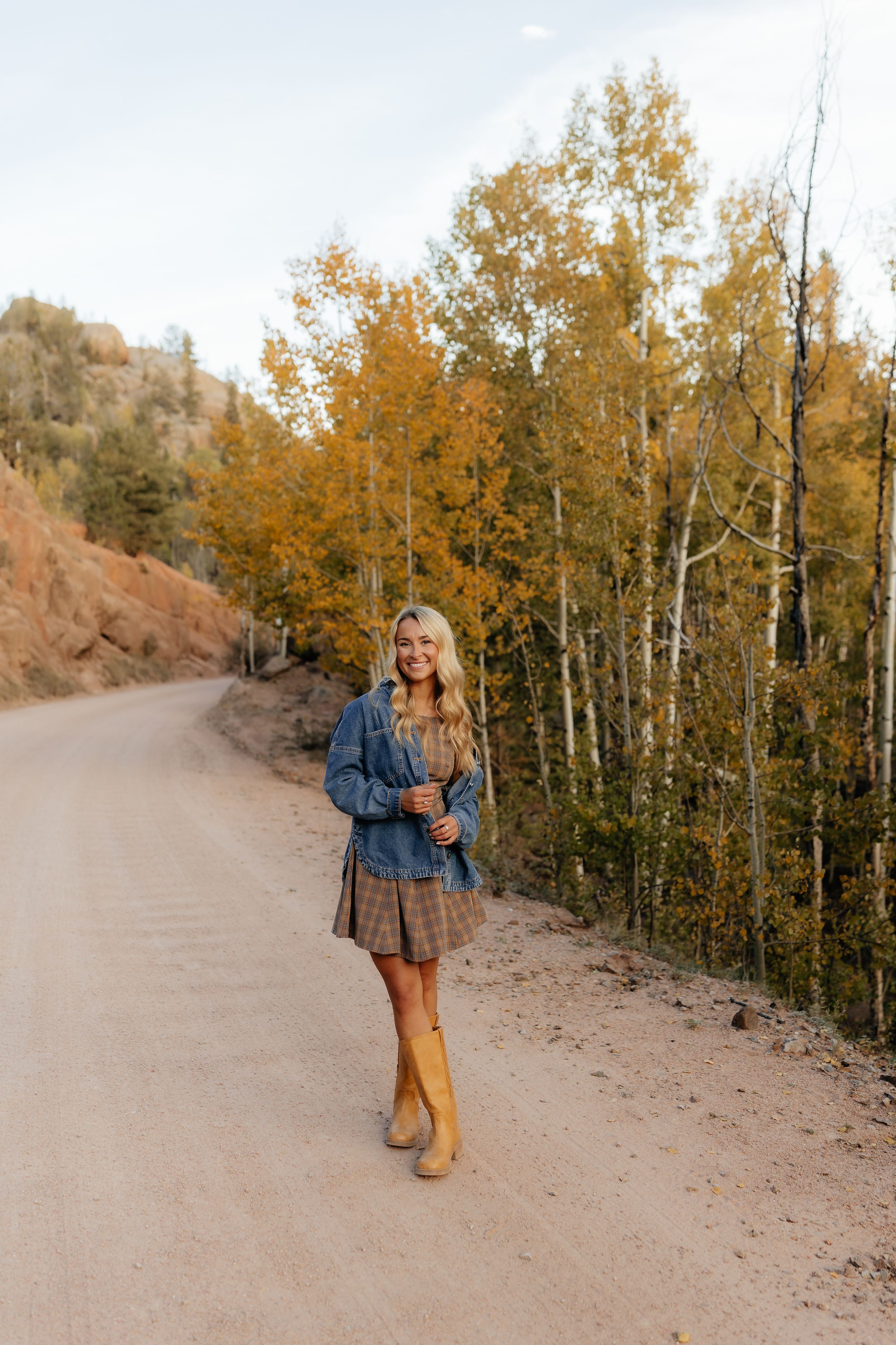 Blonde woman in denim jacket, plaid skirt, and tall boots on a dirt road surrounded by golden autumn aspen trees.